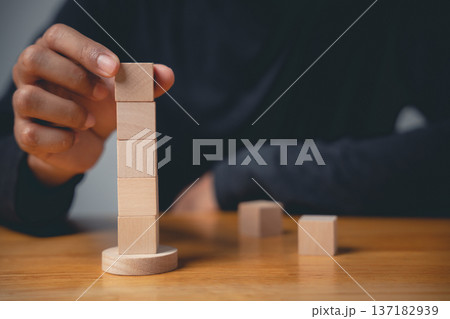 A closeup man hand gesture in a pose builds a tower as a business sign, stacking wooden blocks to create a conceptual sign of progress, growth and development A closeup man hand gesture in a pose builds a tower as a business sign, stacking wooden blocks to create a conceptual sign of progress, growth and development 137182939