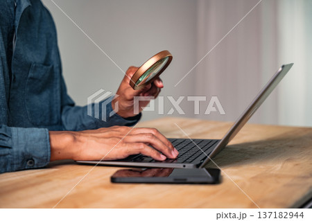 A man in a purposeful business pose holds a magnifier over a laptop, a hand gesture and intense pose that represent concepts like discovery and deep data inspection 137182944