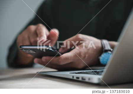 Close up of a person's hands using a smartphone while sitting at a desk with a laptop in the background. 137183010