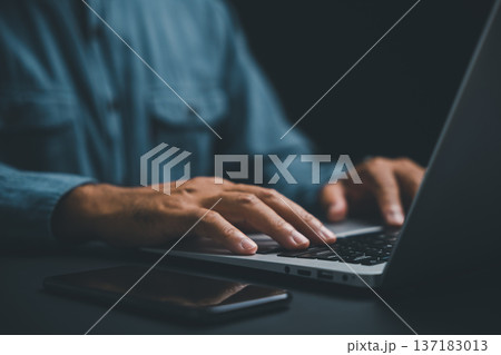 Close-up of hands typing on a laptop keyboard with a smartphone in the foreground. A person's arm and a blue shirt are blurred in the background. Close-up of hands typing on a laptop keyboard with a smartphone in the foreground. A person's arm and a blue shirt are blurred in the background. 137183013