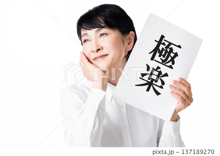 極楽と書かれた紙を持ち頬杖をついて微笑む白いシャツを着たシニア女性 137189270