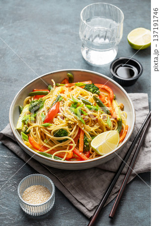 Stir fry wok noodles with vegetables in a plate on a dark background with chopsticks, glass of water Stir fry wok noodles with vegetables in a plate on a dark background with chopsticks, glass of water 137191746
