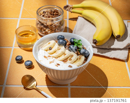 Natural greek yogurt with granola, blueberry and banana in a bowl on a orange tile background Natural greek yogurt with granola, blueberry and banana in a bowl on a orange tile background 137191761