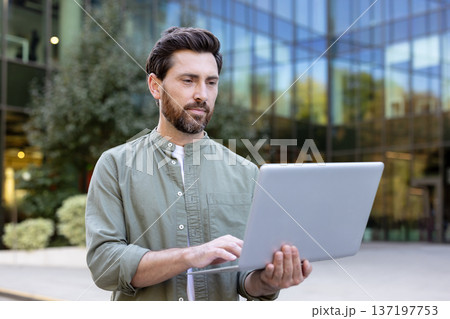 Man standing outdoors in front of a glass building, focused on typing on a laptop, representing remote work, flexible jobs, and mobile business through technology 137197753
