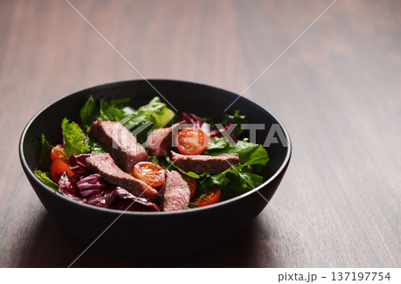 Steak salad with cherry tomatoes and mixed greens in black bowl on walnut wood background 137197754