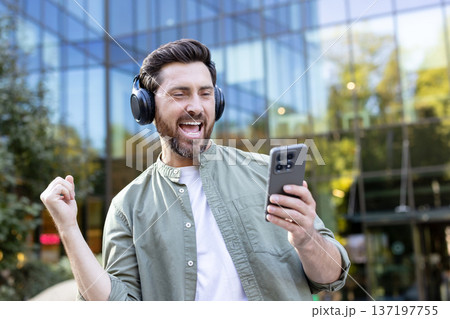 Man celebrating success and experiencing happiness while loudly singing along to music from his smartphone, wearing headphones in an urban environment Man celebrating success and experiencing happiness while loudly singing along to music from his smartphone, wearing headphones in an urban environment 137197755