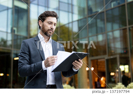 Young professional businessman in a sleek suit carefully reviewing reports and documents on a clipboard while standing outside a contemporary glass corporate building Young professional businessman in a sleek suit carefully reviewing reports and documents on a clipboard while standing outside a contemporary glass corporate building 137197794