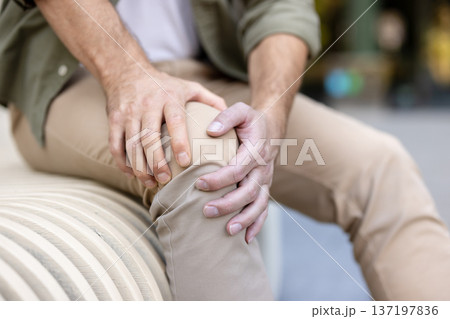 Man crouching outdoors, clutching aching knee in both hands, showing joint pain and discomfort from injury, arthritis or strain, partial close-up, casual clothing, adult male 137197836
