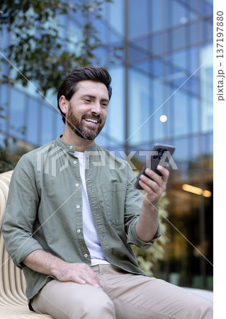 Happy bearded man smiling, engaging in a video call or browsing on his mobile phone while sitting casually on a contemporary bench outside a corporate glass building 137197880