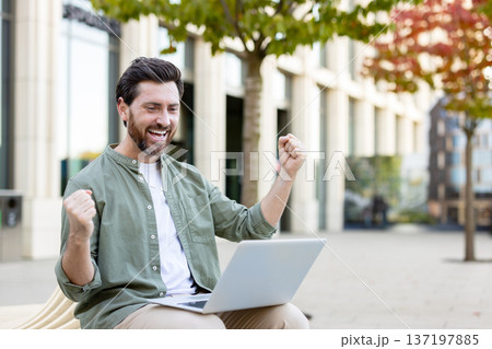 Excited man cheering success while looking at his laptop, sitting on a bench in an urban outdoor setting, personifying achievement, victory, and job well-done Excited man cheering success while looking at his laptop, sitting on a bench in an urban outdoor setting, personifying achievement, victory, and job well-done 137197885