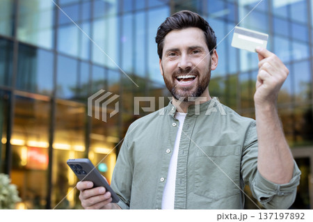 Happy man holding a blank credit card and smartphone, smiling while making an electronic payment or online purchase against a modern blurred building background 137197892