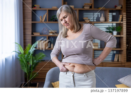 Elderly woman examines stomach with concerned expression. Emphasis on health, wellness, aging, and self-reflection. Scene captures thoughtful awareness of body changes and their emotional impact. 137197899
