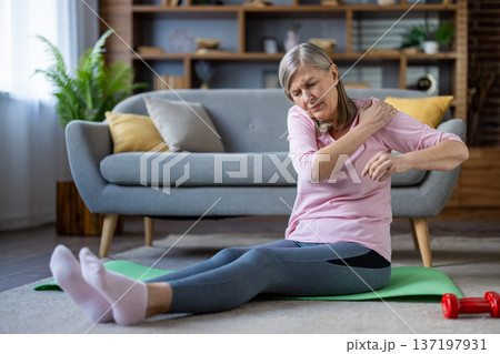 Senior woman with shoulder pain sits on yoga mat in living room. Elderly lady in pink shirt and yoga pants feels discomfort after workout, illustrating challenges of senior fitness. Senior woman with shoulder pain sits on yoga mat in living room. Elderly lady in pink shirt and yoga pants feels discomfort after workout, illustrating challenges of senior fitness. 137197931