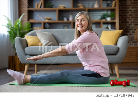 Smiling mature woman doing yoga stretch on mat indoors. Wearing comfortable activewear, she enjoys workout routine for fitness and wellness. Scene captures healthy lifestyle focus in relaxing space. 137197945