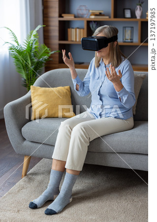 Middle-aged woman using VR headset on couch in modern living room. Experiencing virtual reality, exploring immersive technology with curiosity and excitement in casual setting. Lifestyle concept. 137197985