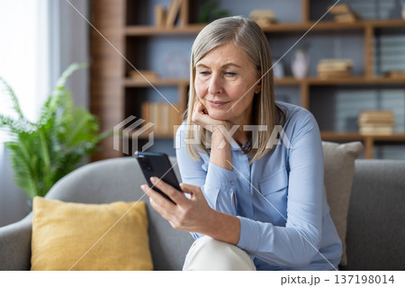 Senior woman with gray hair using smartphone, sitting on sofa, looking thoughtful. She appears relaxed and engaged with technology, possibly checking messages or social media on phone. 137198014
