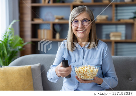 Smiling elderly woman holding bowl of popcorn and remote, sitting on cozy sofa. Leisure time enjoyment at home, ready for watching TV. Comfortable relaxed setting conveys happiness. Smiling elderly woman holding bowl of popcorn and remote, sitting on cozy sofa. Leisure time enjoyment at home, ready for watching TV. Comfortable relaxed setting conveys happiness. 137198037