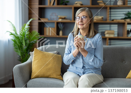Senior woman sitting on couch holding coffee mug. Woman enjoying peaceful moment at home, feeling relaxed and content. Light-filled living room with stylish decor in background 137198053