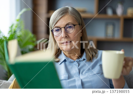 Senior woman wearing glasses reading book while drinking coffee at home. Relaxed elderly lady enjoying quiet time and leisure activity in cozy living room environment. Senior woman wearing glasses reading book while drinking coffee at home. Relaxed elderly lady enjoying quiet time and leisure activity in cozy living room environment. 137198055