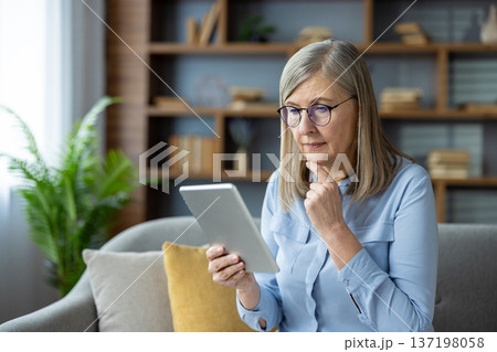 Senior woman with glasses reading on tablet in modern living room thoughtfully. She seems deeply engaged with information on the device showing concentration and curiosity. 137198058