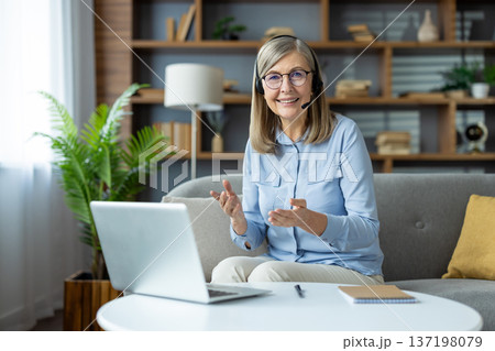 Senior woman communicating with clients through video call on laptop at home, smiling and using headset. Comfortable home office setup with plant, books, and notepad on the table Senior woman communicating with clients through video call on laptop at home, smiling and using headset. Comfortable home office setup with plant, books, and notepad on the table 137198079