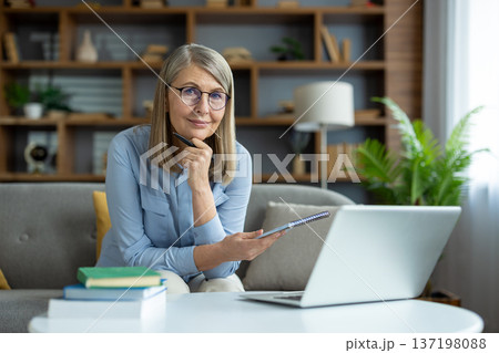 Mature woman working from home on laptop, engaging in online teaching or virtual meeting. Sitting on couch, holding notebook, and pen. Comfortable home office environment with books and plants. Mature woman working from home on laptop, engaging in online teaching or virtual meeting. Sitting on couch, holding notebook, and pen. Comfortable home office environment with books and plants. 137198088