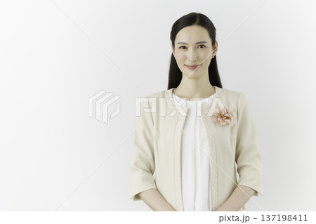 白背景の白いブラウスにクリーム色のジャケットを着て胸に花のコサージュをつけて微笑む若い日本人女性 137198411