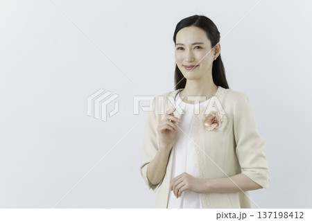 白背景の白いブラウスにクリーム色のジャケットを着て胸に花のコサージュをつけて微笑む若い日本人女性 137198412