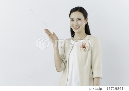 白背景の白いブラウスにクリーム色のジャケットを着て胸に花のコサージュをつけて手をかざす女性 137198414
