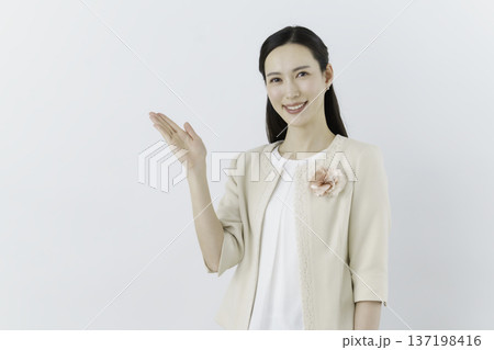 白背景の白いブラウスにクリーム色のジャケットを着て胸に花のコサージュをつけて手をかざす女性 白背景の白いブラウスにクリーム色のジャケットを着て胸に花のコサージュをつけて手をかざす女性 137198416