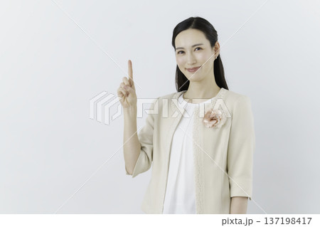 白背景の白いブラウスにクリーム色のジャケットを着て胸に花のコサージュをつけて1番のポーズをする女性 白背景の白いブラウスにクリーム色のジャケットを着て胸に花のコサージュをつけて1番のポーズをする女性 137198417