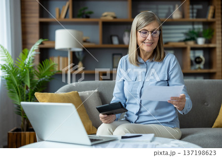 Senior woman sitting on couch, using laptop and reviewing financial documents. Concept of home finances, budget management, and senior independent living. 137198623