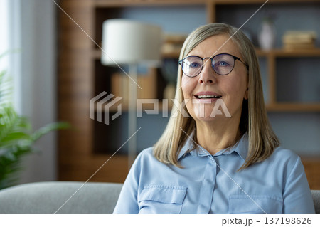 Smiling mature woman with glasses looking happy while sitting in cozy living room. Modern home interior with bookshelf and lamp in background Smiling mature woman with glasses looking happy while sitting in cozy living room. Modern home interior with bookshelf and lamp in background 137198626