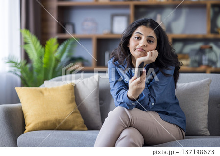 Portrait of a bored Indian young girl sitting on the sofa at home and looking at the camera, leaning on her hand. 137198673