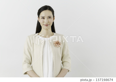 白背景の白いブラウスにクリーム色のジャケットを着て胸に花のコサージュをつけて微笑む若い日本人女性 137198674