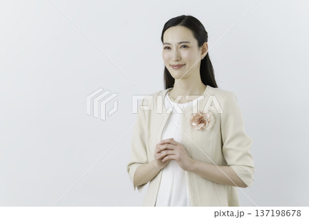白背景の白いブラウスにクリーム色のジャケットを着て胸に花のコサージュをつけて手を組む女性 137198678