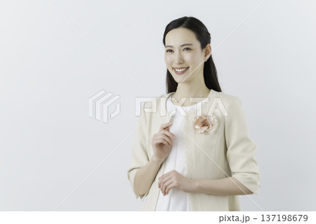 白背景の白いブラウスにクリーム色のジャケットを着て胸に花のコサージュをつけて手をかざす女性 白背景の白いブラウスにクリーム色のジャケットを着て胸に花のコサージュをつけて手をかざす女性 137198679