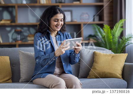 A smiling woman sits comfortably on a sofa, focused on her tablet. She appears relaxed in her living room setting, engaging with her device. 137198816