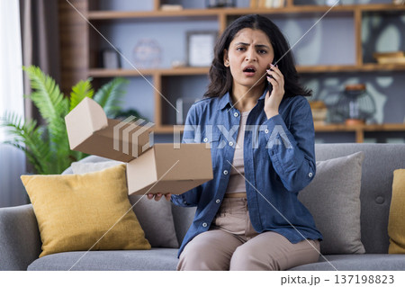 Upset young Indian woman is at home, sitting on the sofa, holding an open box in her hands and talking emotionally on the phone. Upset young Indian woman is at home, sitting on the sofa, holding an open box in her hands and talking emotionally on the phone. 137198823