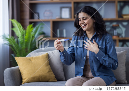 Happy young Indian woman sitting on the couch at home and rejoicing at the news of pregnancy, holding and looking at a disposable test in her hands. Happy young Indian woman sitting on the couch at home and rejoicing at the news of pregnancy, holding and looking at a disposable test in her hands. 137198845