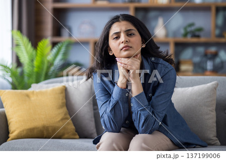 Portrait of a sad and worried young Muslim woman sitting on the couch at home, folding her hands together and looking sadly at the camera. Portrait of a sad and worried young Muslim woman sitting on the couch at home, folding her hands together and looking sadly at the camera. 137199006