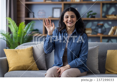 Portrait of a young smiling Indian woman in casual outfit sitting on the couch at home, waving and greeting the camera. Portrait of a young smiling Indian woman in casual outfit sitting on the couch at home, waving and greeting the camera. 137199033