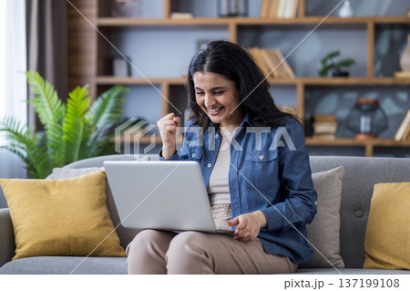 Happy Indian young woman sitting on the sofa at home, holding a laptop on her lap and rejoicing in success, showing a victory gesture. Happy Indian young woman sitting on the sofa at home, holding a laptop on her lap and rejoicing in success, showing a victory gesture. 137199108