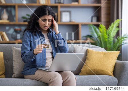 Shocked young Indian girl sitting on the couch at home, holding a laptop on her lap, holding her head in concern and looking at the credit card she is holding in her hand. Shocked young Indian girl sitting on the couch at home, holding a laptop on her lap, holding her head in concern and looking at the credit card she is holding in her hand. 137199244