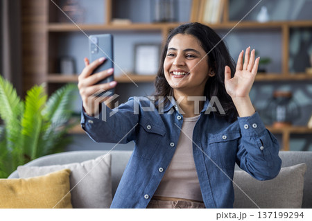 Smiling young Indian woman sitting on sofa at home, talking on video call and greeting on mobile phone camera. 137199294