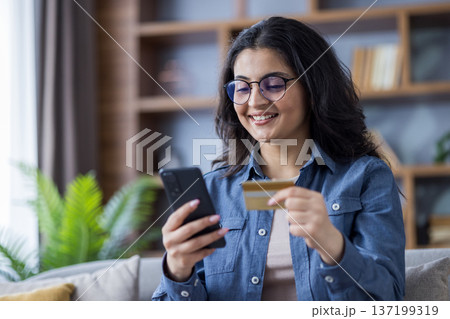 Close-up photo of a young Indian woman sitting on the sofa at home, holding a credit card and using a mobile phone. 137199319