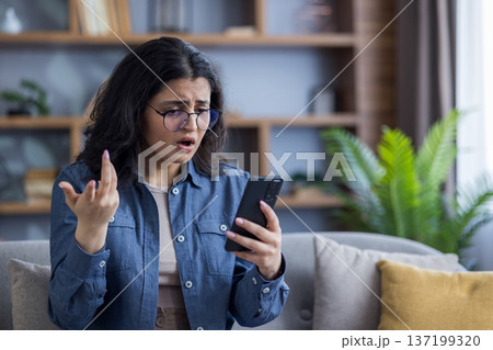 A worried young Indian girl sits at home on the sofa and looks disappointedly at the screen of the mobile phone she is holding in her hand. 137199320