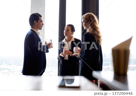 Business professionals networking by a large window at a corporate event, discussing strategy and partnership while holding drinks in a modern office setting with city view background Business professionals networking by a large window at a corporate event, discussing strategy and partnership while holding drinks in a modern office setting with city view background 137200448