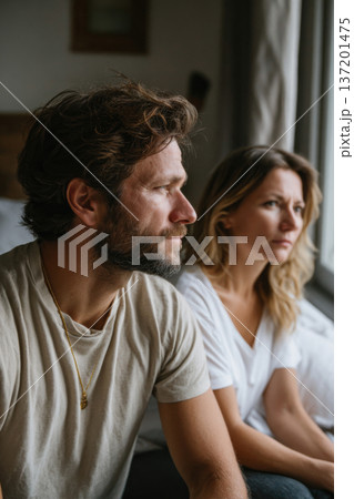 adult couple sitting on bed side by side after argument, faces turned away from camera, soft side window light, neutral bedroom interior 137201475