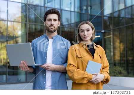 Two professional business people, a man holding a laptop and a woman holding a tablet, standing confidently outside a modern glass office building, representing teamwork and corporate success Two professional business people, a man holding a laptop and a woman holding a tablet, standing confidently outside a modern glass office building, representing teamwork and corporate success 137202721